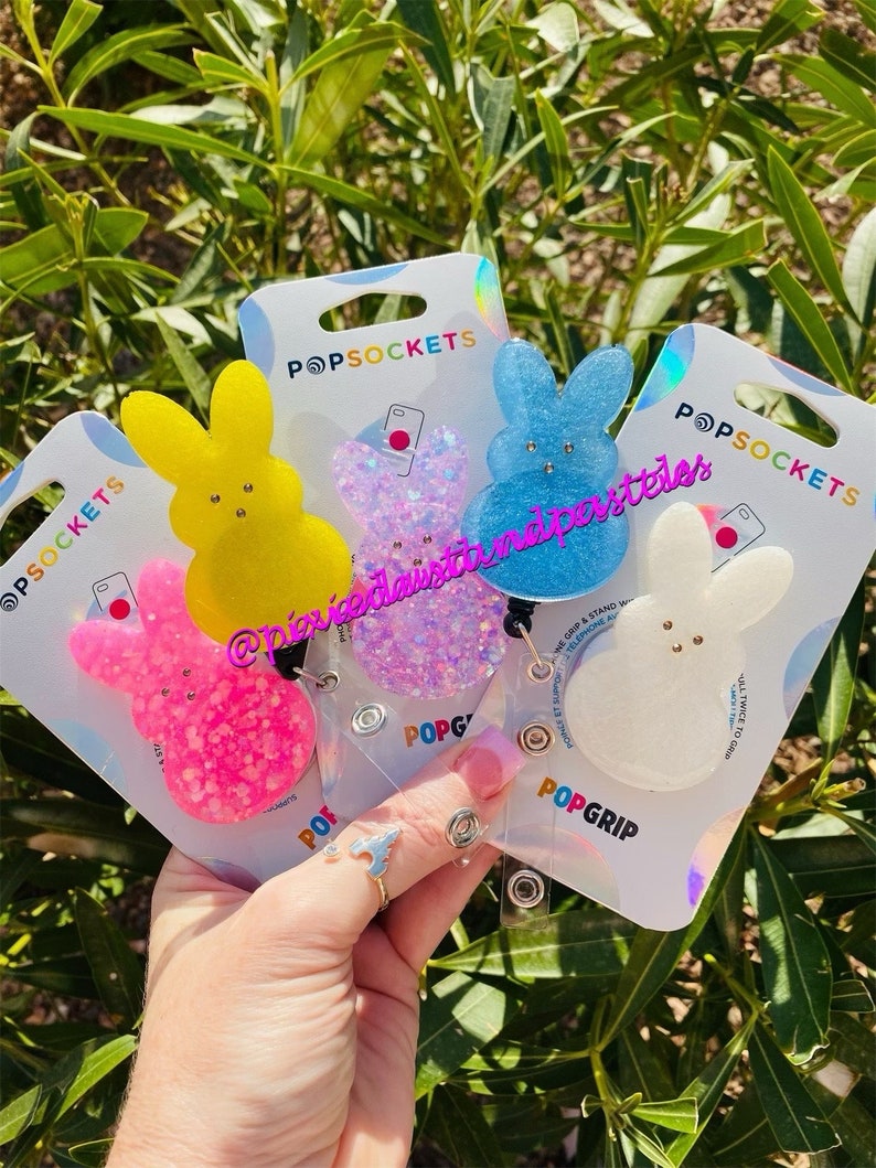 May include: Four colorful, glittery, bunny-shaped phone grips with a white background. The phone grips are attached to clear plastic badge holders with the text "POPGRIP" printed on them. The phone grips are in the colors pink, yellow, blue, and white.