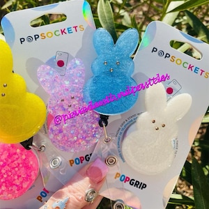 May include: Three PopSockets phone grips featuring a bunny design. The grips are made of clear resin with glitter embedded in the resin. The bunny designs are in pink, blue, and white.