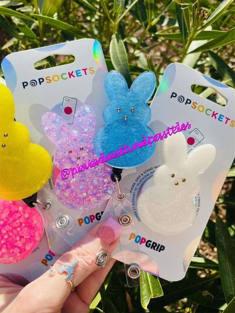 May include: Three PopSockets phone grips featuring a bunny design. The grips are made of clear resin with glitter embedded in the resin. The bunny designs are in pink, blue, and white.