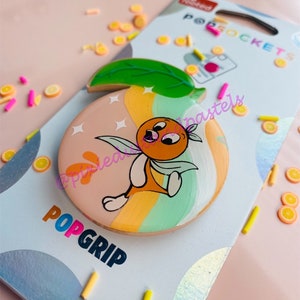 May include: A round orange PopGrip phone grip with a cartoon orange character and a green leaf. The background is white with the words "PopGrip" in rainbow colors.
