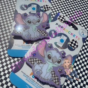 May include: Two PopSockets phone grips featuring the character Stitch from Disney's Lilo & Stitch. The grips are light blue, purple, and white, with a checkered pattern. The packaging includes the PopSockets logo and the Disney logo.