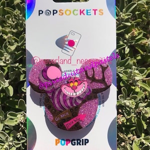 May include: A pink and purple glitter phone grip featuring the Cheshire Cat from Alice in Wonderland. The grip has a pop-out feature and says "Go Back This Way" on the back.