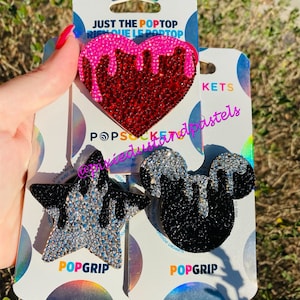 May include: Three phone grip designs with rhinestones. A red heart with pink dripping, a black Mickey Mouse head with black dripping, and a silver star with silver dripping. Each design has the text "POPGRIP" below it.