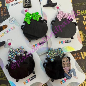 May include: A collection of black PopSockets featuring cauldron designs with various embellishments, including green, pink, and iridescent gems. Other designs include a witch hat and a miniature book. The PopSockets are displayed on branded packaging.