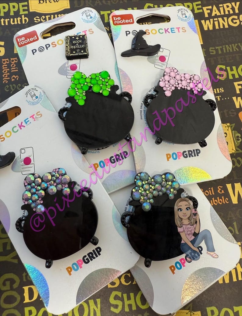 May include: A collection of black PopSockets featuring cauldron designs with various embellishments, including green, pink, and iridescent gems. Other designs include a witch hat and a miniature book. The PopSockets are displayed on branded packaging.