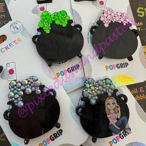 May include: A collection of black PopSockets featuring cauldron designs with various embellishments. Some have green, pink, or iridescent crystal-like accents, and one includes a witch hat. The PopSockets are on branded packaging.
