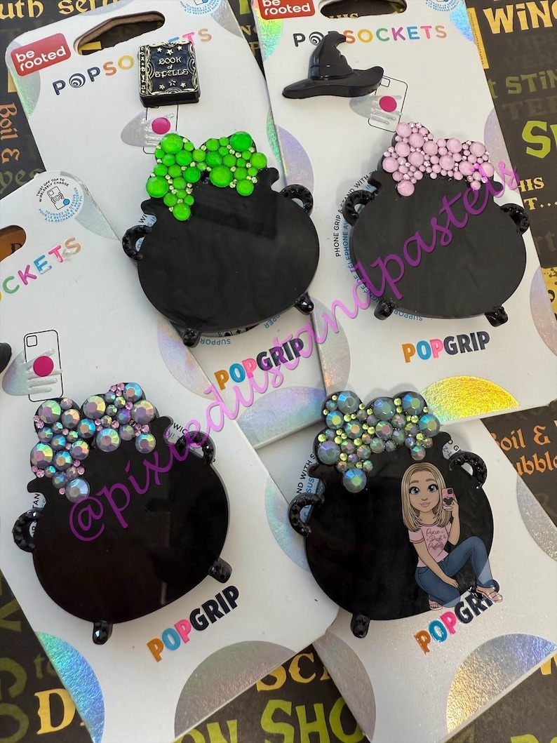 May include: A collection of black PopSockets featuring cauldron designs with various embellishments. Some have green, pink, or iridescent crystal-like accents, and one includes a witch hat. The PopSockets are on branded packaging.