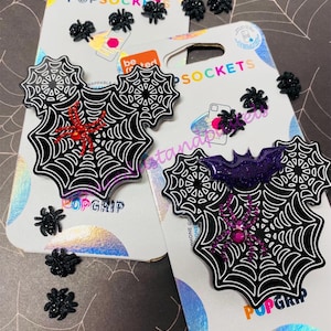 Op de afbeelding: Twee zwarte en witte telefoonbeugels in de vorm van een spinnenweb met een paarse en rode glitterspin op elk. De beugels bevinden zich op een verpakking met de tekst "PopSockets" en "PopGrip".
