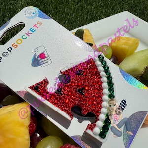 May include: A PopSockets phone grip featuring a watermelon slice design, covered in red rhinestones with black seed accents. The grip has a white border and a green rhinestone rind. The packaging is visible.
