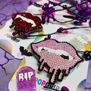 Puede incluir: Tres agarres para teléfono con temática de Halloween. Uno es un labio de vampiro rosa y blanco que gotea con detalles negros. Otro es un labio de vampiro rojo con colmillos blancos. El tercero es una lápida morada con la palabra "RIP" en blanco.