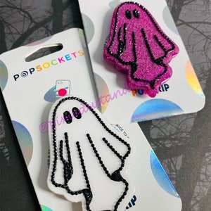 May include: Two PopSockets phone grips featuring a ghost design. One grip is white with black glitter and the other is pink with black glitter.