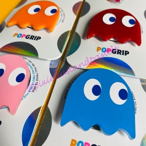 May include: Four phone grips featuring a Pac-Man ghost design. The grips are in pink, orange, red, and blue. Each grip has the text "POPGRIP" printed on the bottom.