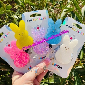 May include: Four PopSockets phone grips featuring a bunny design. The grips are made of glitter and come in pink, blue, white, and yellow. The grips are attached to clear plastic badge holders with a retractable cord.