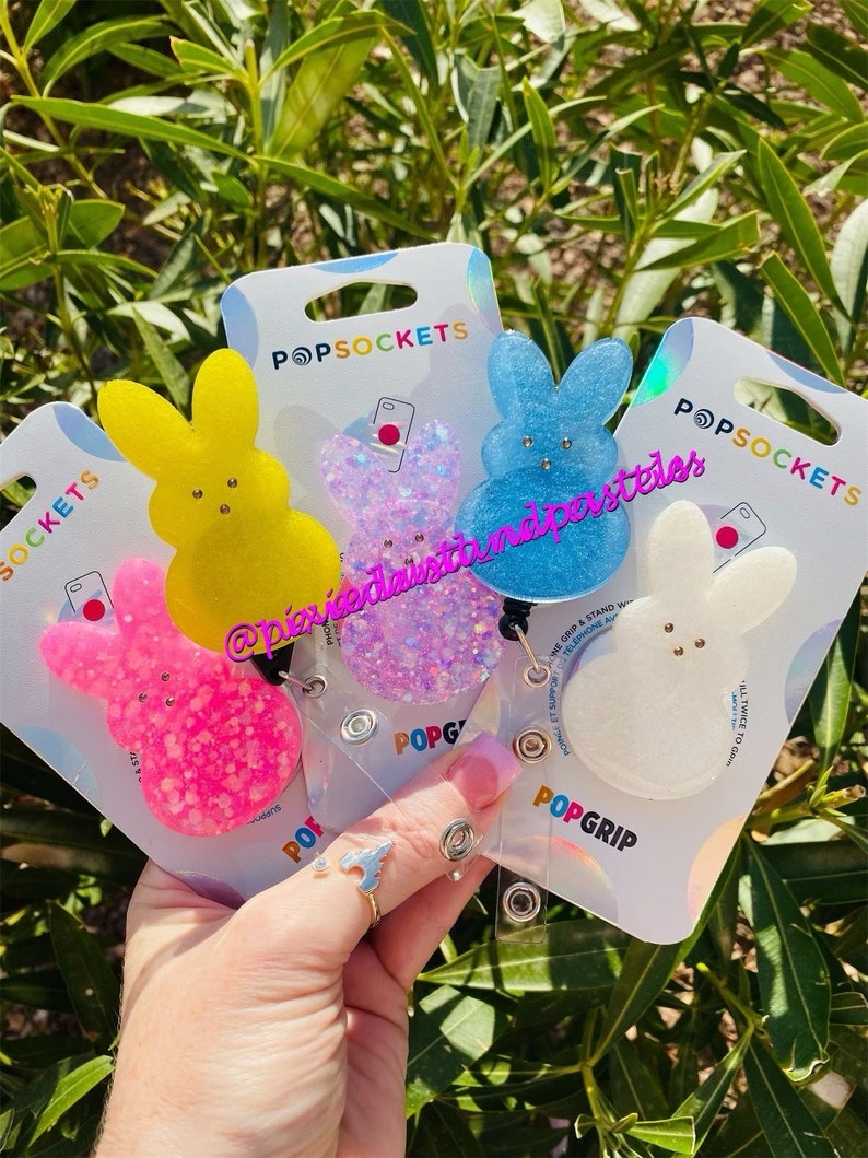 May include: Four PopSockets phone grips featuring a bunny design. The grips are made of glitter and come in pink, blue, white, and yellow. The grips are attached to clear plastic badge holders with a retractable cord.