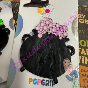 May include: A black PopSocket with a witch's cauldron design. The cauldron is black with pink crystal accents and a small black witch hat. The PopSocket is on a white backing with the word "POPGRIP" in colorful letters.