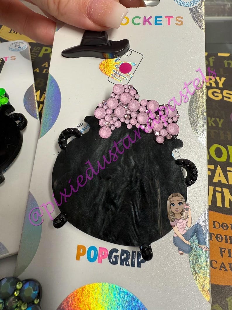 May include: A black PopSocket with a witch's cauldron design. The cauldron is black with pink crystal accents and a small black witch hat. The PopSocket is on a white backing with the word "POPGRIP" in colorful letters.
