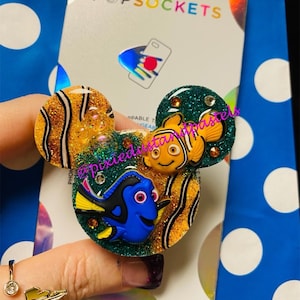 May include: A PopSockets phone grip featuring a green, orange, and yellow glitter background with a blue and yellow Dory and an orange and yellow Nemo from the movie Finding Nemo.