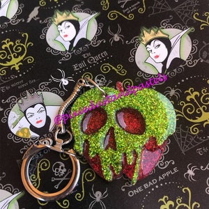 May include: A green and red glitter-covered apple keychain with a skull design. The apple is dripping with red glitter and has the text "One Bad Apple" on the bottom. The keychain is attached to a silver ring.