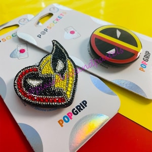 May include: Two phone grips with a black, red, and yellow design. One grip features a Deadpool logo and the other features a Wolverine logo. Both grips are on white packaging with the text "POPGRIP" and "POPSOCKETS".