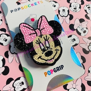 Pode incluir: Um agarre para telefone PopSocket Minnie Mouse preto e rosa com acabamento brilhante. O PopSocket está em um cartão branco com as palavras "PopSockets" e "PopGrip" impressas nele. O fundo é um padrão rosa de rostos de Minnie Mouse.