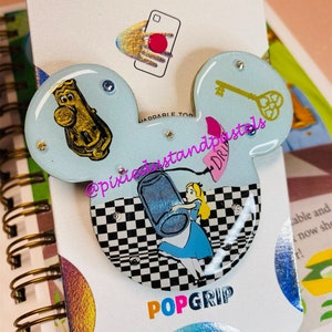 May include: A blue and white PopGrip phone accessory with a checkered background. The PopGrip features a depiction of Alice from Alice in Wonderland holding a bottle. The PopGrip is attached to a blue Mickey Mouse ear-shaped phone stand with a gold key and a golden figure of Mr. Potato Head.