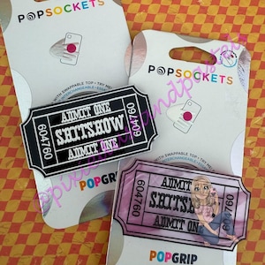Pode incluir: Dois suportes de telefone PopSockets com designs de bilhetes. Um bilhete preto e branco que diz "ADMIT ONE SHITSHOW". O outro é rosa e apresenta uma menina de desenho animado. Ambos têm o número 604760.