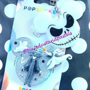 May include: A white, glittery phone grip with a Jack Skellington and Zero character design from the Nightmare Before Christmas movie. The grip is attached to a silver keychain. The packaging includes the text "PopSocket" and "PopGrip".