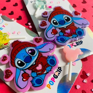 May include: Two Stitch character pop grips with a heart-shaped lollipop. The Stitch characters are wearing red sweaters and have blue fur. The pop grips are purple and pink.