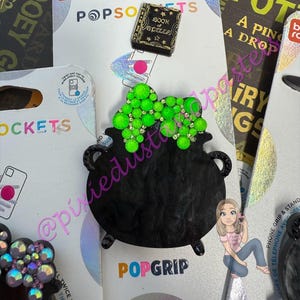 May include: Black cauldron PopGrip with green gem accents, a 'Book of Spells' charm, and a PopSockets package. The PopGrip is designed to attach to the back of a phone for a secure grip and stand.