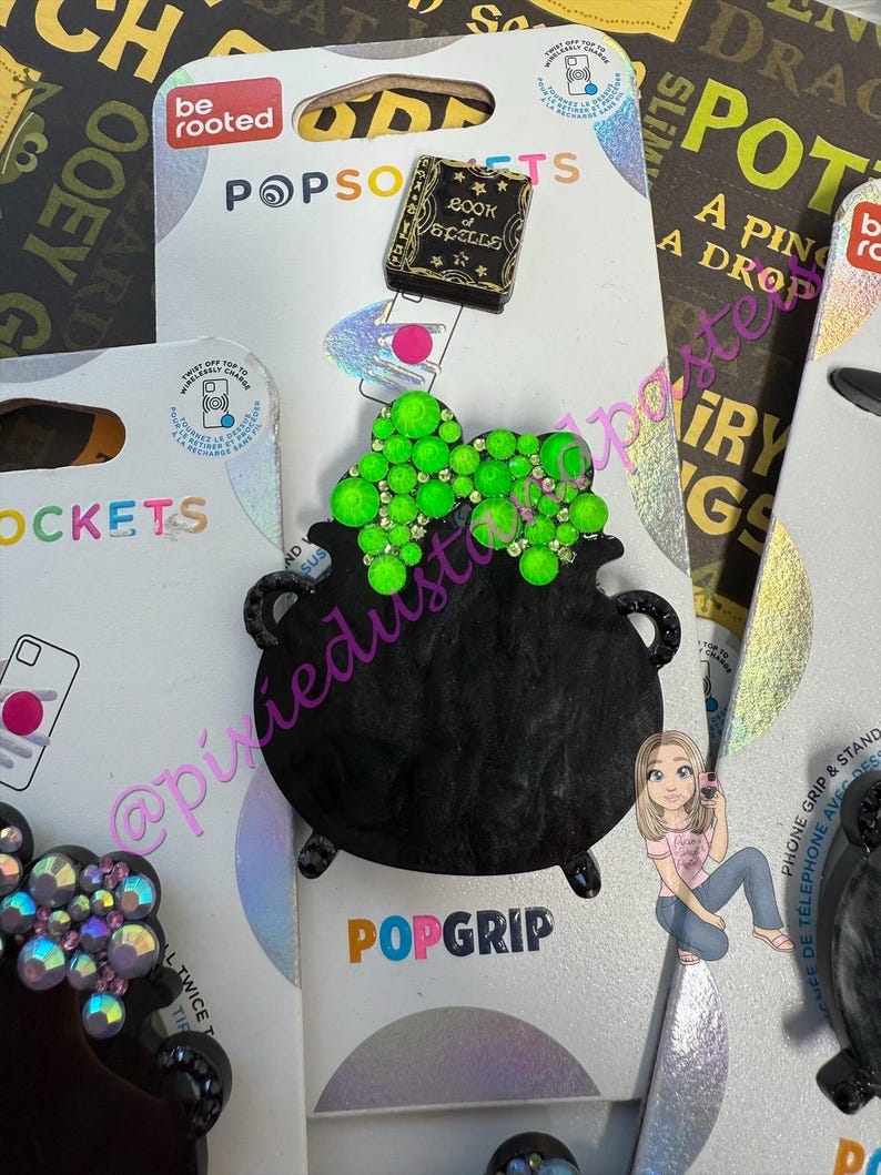 May include: Black cauldron PopGrip with green gem accents, a 'Book of Spells' charm, and a PopSockets package. The PopGrip is designed to attach to the back of a phone for a secure grip and stand.
