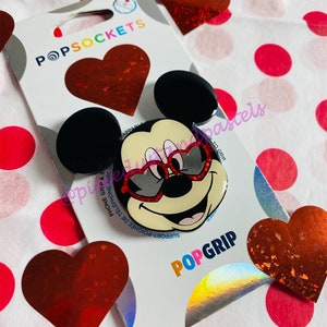 May include: A red and black PopSockets phone grip featuring a cartoon Mickey Mouse wearing red sunglasses. The background is a white surface with red polka dots.