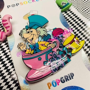 May include: A pink teacup with a Mad Hatter character from Alice in Wonderland riding inside. The teacup is on a white background with the words "PopSockets" and "PopGrip" in colorful letters.