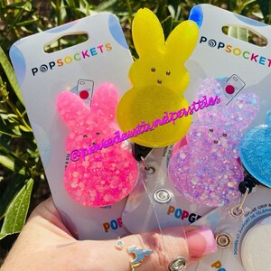 May include: Three colorful phone grips shaped like Peeps, a popular marshmallow candy. The grips are pink, yellow, and blue with glitter. The packaging includes the text "POPSOCKETS" and "POPGRIP".