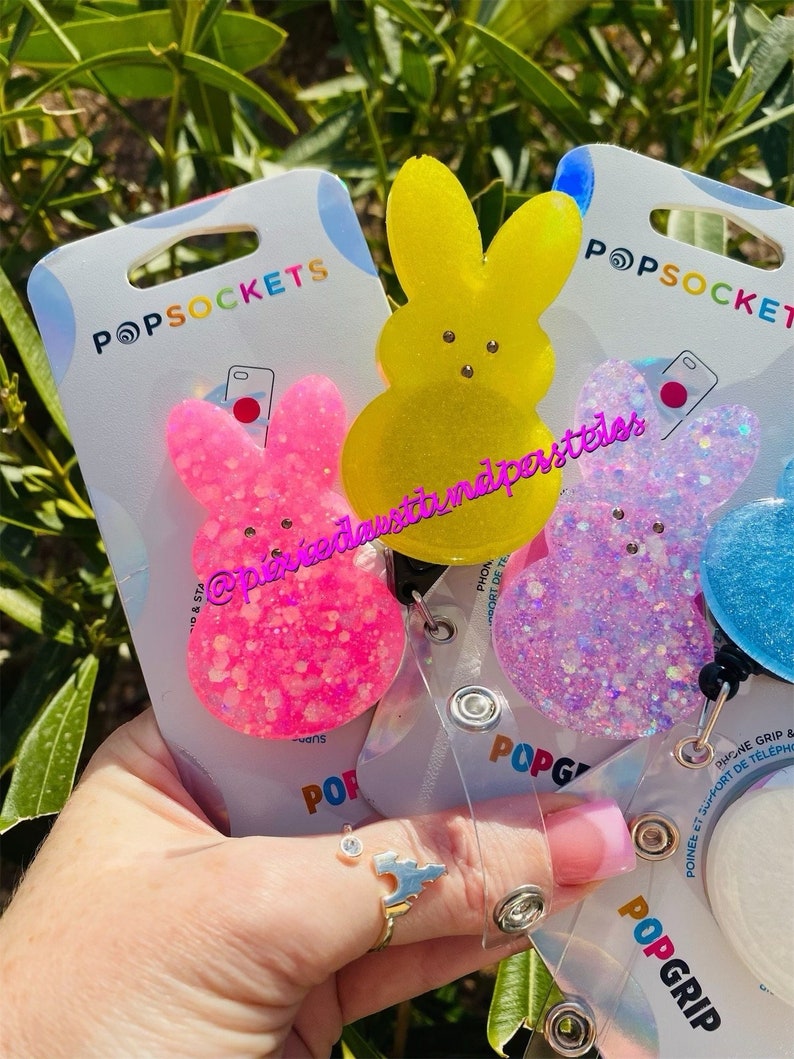 May include: Three colorful phone grips shaped like Peeps, a popular marshmallow candy. The grips are pink, yellow, and blue with glitter. The packaging includes the text "POPSOCKETS" and "POPGRIP".