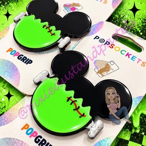 Könnte beinhalten: Zwei PopSockets PopGrip Handyzubehör mit einer schwarzen Mickey-Maus-Kopf-Silhouette, einem leuchtend grünen Unterteil, roten Nahtdetails und silberfarbenen Bolzen an den Seiten. Die Verpackung zeigt den Markennamen PopSockets.