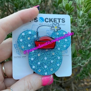 May include: A blue and white glitter phone grip with a red and white bucket and a white flower. The grip is shaped like Mickey Mouse ears. The text "SOCKETS" is on the card behind the grip. The text "USE YOUR PHONE BETTER" is also on the card.