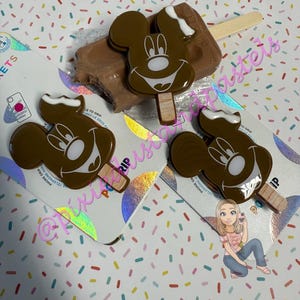 Könnte beinhalten: Braune Mickey Mouse-förmige Telefonhalterungen mit weißen Akzenten und Holzstäbchen, die Eis am Stiel ähneln. Ein echter Eisriegel ist ebenfalls vorhanden. Die Halterungen befinden sich auf schillernden Karten mit dem Text "PHONE GRIP". Der Hintergrund ist ein Konfetti-Muster.