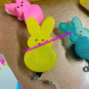 May include: Three pastel-colored bunny-shaped keychains with glitter. The keychains are yellow, pink, and blue. Each keychain has two black dots for eyes.