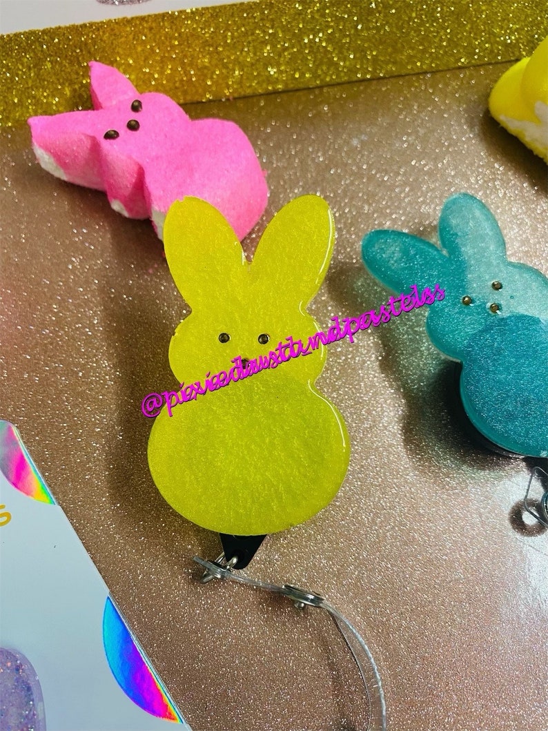May include: Three pastel-colored bunny-shaped keychains with glitter. The keychains are yellow, pink, and blue. Each keychain has two black dots for eyes.