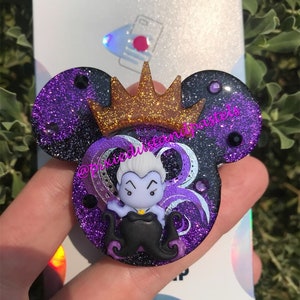 May include: Purple and black glitter phone grip with a gold crown and a cartoon image of Ursula from The Little Mermaid. The grip is shaped like Mickey Mouse ears.