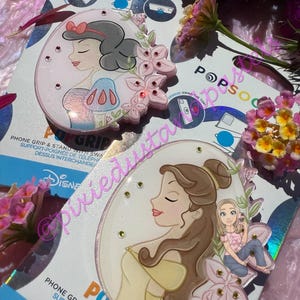 May include: Two PopSockets phone grips featuring Disney princesses Snow White and Belle. Each grip has a pink floral design with rhinestones and a portrait of the princess. The packaging includes the PopSocket logo and product information.