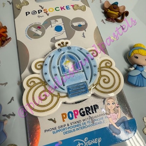 May include: A PopSocket phone grip with a light blue Cinderella carriage design and gold detailing. The packaging features the PopSocket and Disney branding. The grip is designed to attach to the back of a phone.