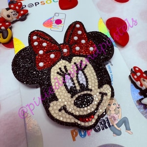 May include: A black and red Minnie Mouse PopSockets phone grip with a red bow and white polka dots. The face is cream-colored and embellished with rhinestones. The PopSocket is on a white backing with the PopSockets logo.