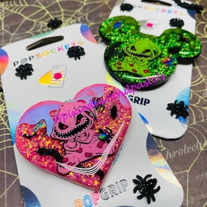 Könnte beinhalten: Zwei Handygriffe mit einem grünen und pinkfarbenen Charakter mit einem genähten Aussehen. Die Griffe sind mit Glitzer und schwarzen Spinnen-Akzenten verziert. Die Verpackung enthält den Text "PopSockets" und "PopGrip".