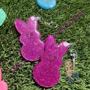 May include: Two pink glitter bunny-shaped keychains with silver key rings. The keychains are on a green grass background.