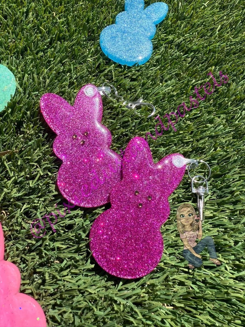 May include: Two pink glitter bunny-shaped keychains with silver key rings. The keychains are on a green grass background.
