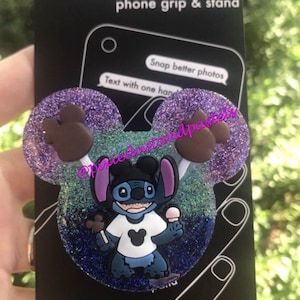 Puede incluir: Una agarradera para teléfono y un soporte de purpurina morada y azul con un diseño de oreja de Mickey Mouse marrón. El centro presenta un personaje azul Stitch sosteniendo un cono de helado de Mickey Mouse y un pretzel de Mickey Mouse. El texto "Popsckets phone grip & stand" está en el empaque.