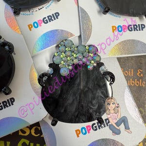 May include: Black PopGrip phone grip featuring a cauldron design with iridescent rhinestones. The PopGrip has a black base and a holographic backing. The word "POPGRIP" is printed in orange, pink, and blue.