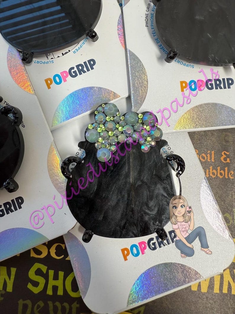 May include: Black PopGrip phone grip featuring a cauldron design with iridescent rhinestones. The PopGrip has a black base and a holographic backing. The word "POPGRIP" is printed in orange, pink, and blue.