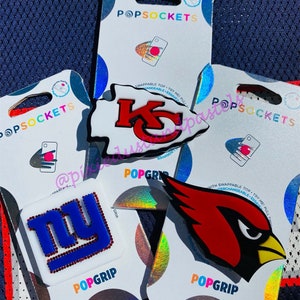 May include: Four PopSockets phone grips with interchangeable tops. The tops feature NFL team logos: Kansas City Chiefs, New York Giants, Arizona Cardinals, and a generic football.